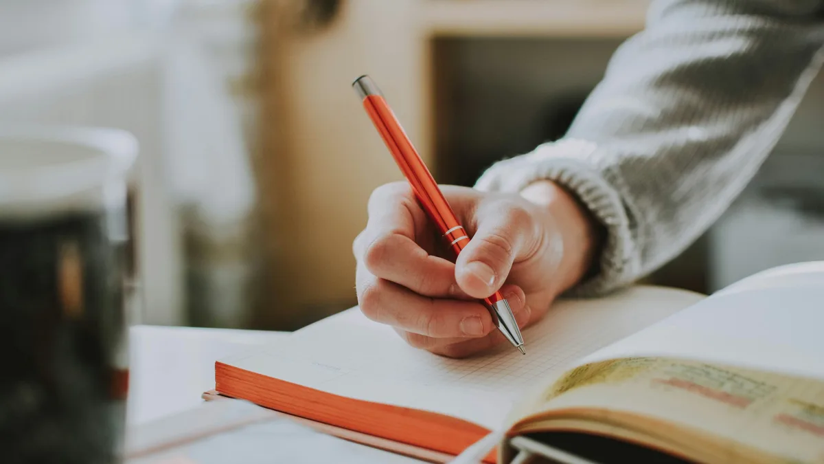 Uma mão segurando uma caneta vermelha e escrevendo em um caderno aberto