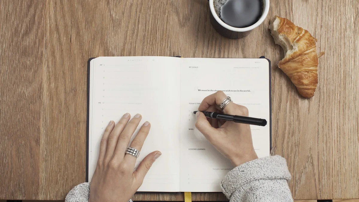 Hands writing in an open journal next to a cup of coffee and a croissant
